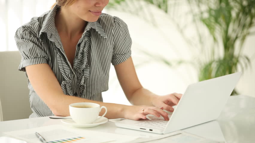 Pretty girl sitting at office table with cup of tea using laptop looking at