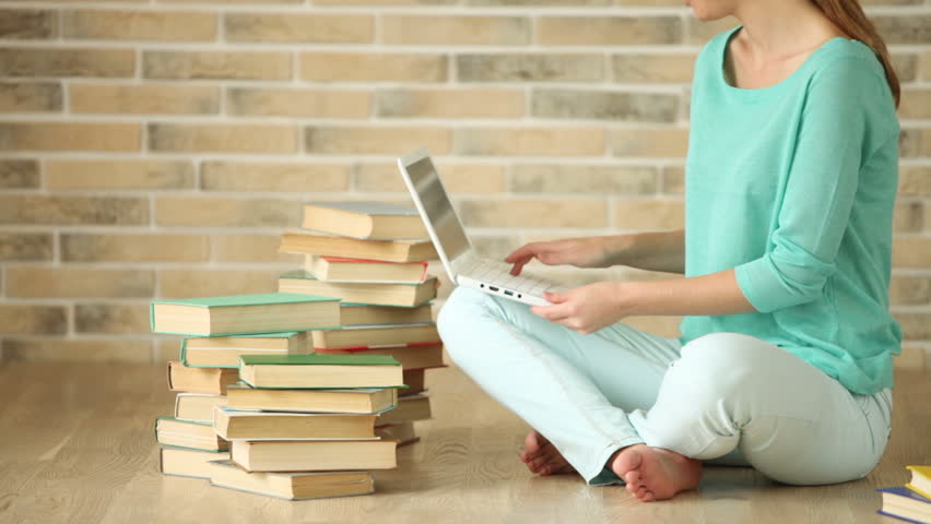 Cute girl sitting on floor with books using laptop looking at camera and