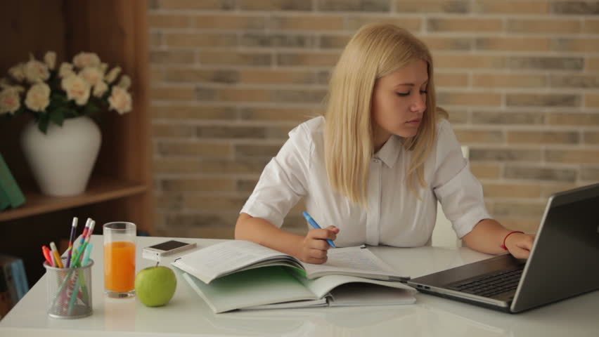 Cheerful girl sitting at desk writing in notebook using laptop looking at camera