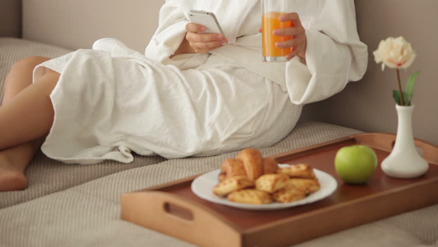 Cheerful girl in bathrobe sitting on sofa with tray of food using cellphone