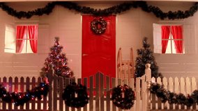 Slow motion panorama video of a red front door of the white house nicely decorated for Christmas with fir trees, a wreath and with sparkling lights.