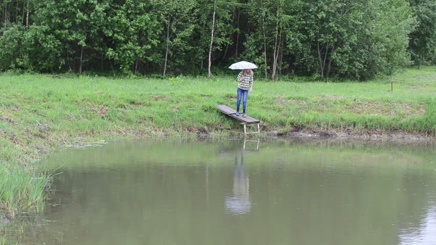 Farm woman with umbrella stand on wooden pond bridge in rain and show fishes. Fish breeding.