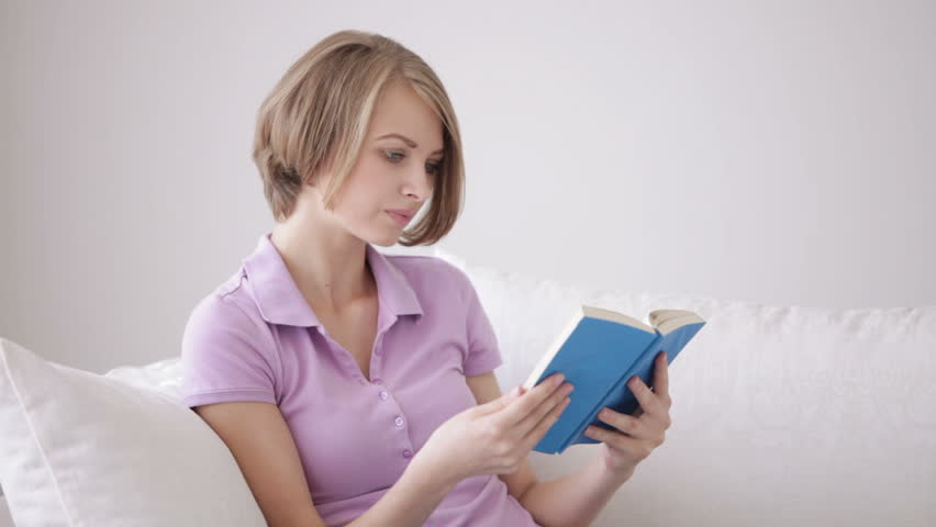 Pretty young woman sitting on sofa reading book looking at camera and smiling