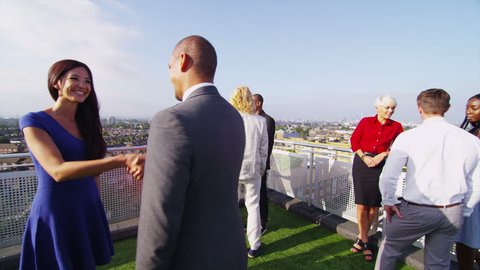 Smartly dressed group of friends or business colleagues chatting outdoors on a rooftop terrace overlooking the city. One man and woman meet and shake hands.