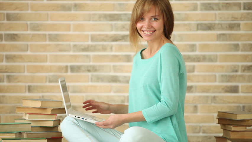 Attractive girl sitting on floor with books using laptop looking at camera and