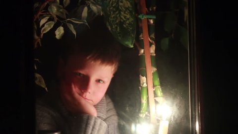 A boy looks out the window at night, lit candle on a windowsill
