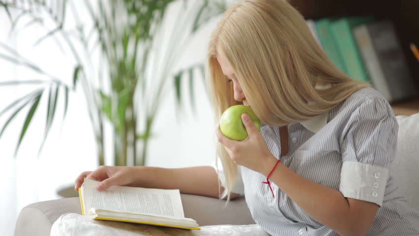 Cute girl sitting on sofa reading book and eating apple. Panning camera