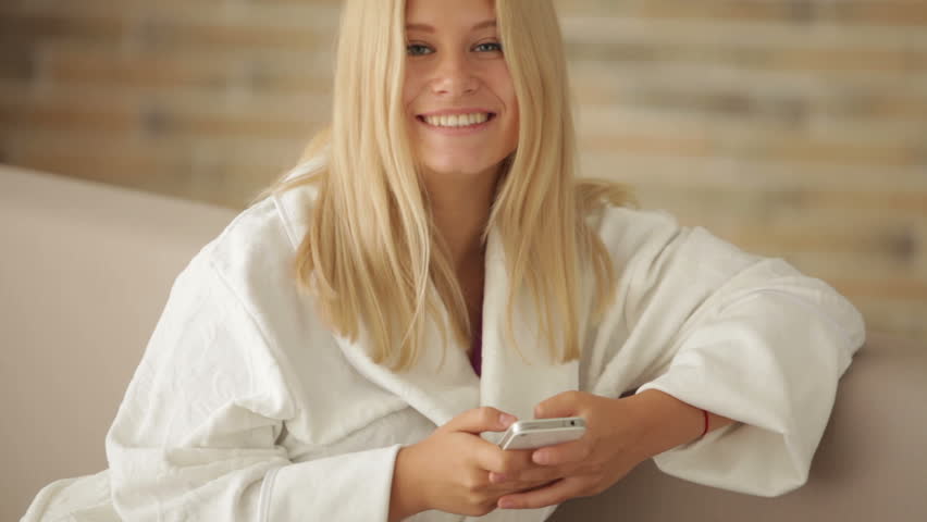 Young woman in bathrobe relaxing on sofa using cellphone looking at camera and