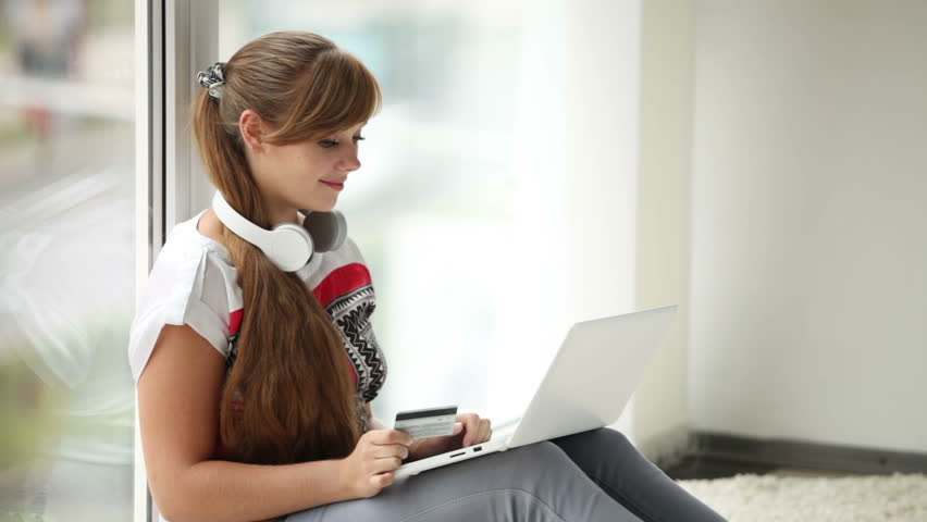 Attractive girl in headset sitting by window using laptop holding credit card
