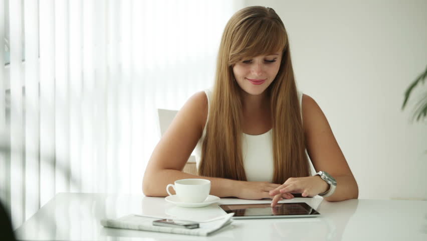 Beautiful girl sitting at table drinking coffee using touchpad looking at camera