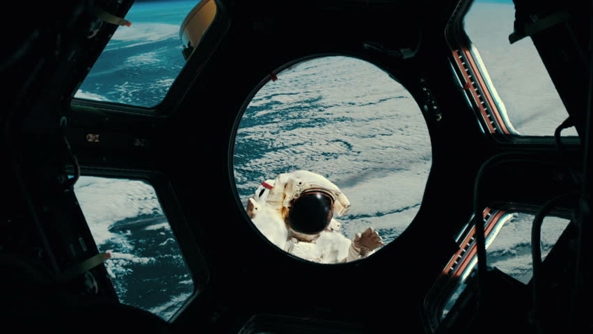 Astronaut waves to other astronauts through space station window.