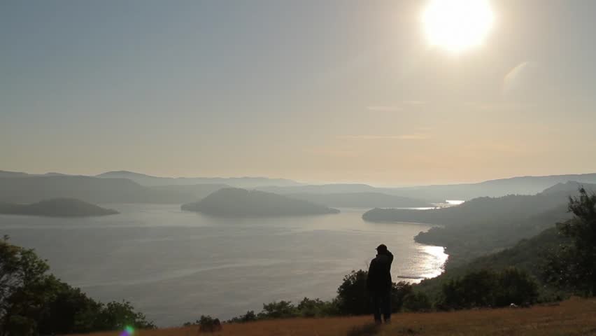 Sunset over giant mountain lake with island. Woman silhouette standing and watching to the sun and water.
