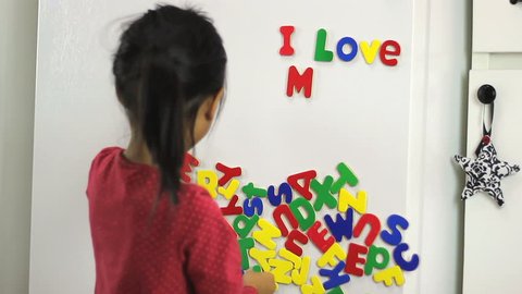 A cute little seven year old Asian girl uses colorful fridge magnet letters to spell I Love Mom on the fridge.