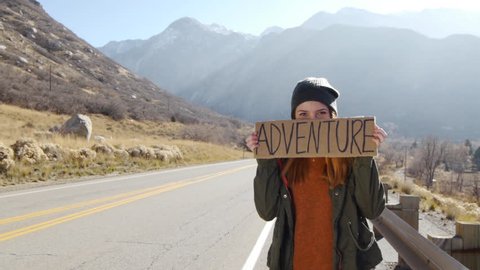 Teenage Girl Holding An Adventure Sign On A Mountain Road, Smiles And Laughs