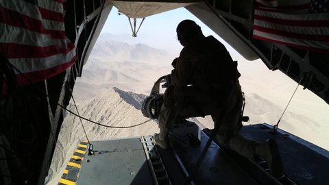 Tail Gunner Sits in Open Doorway of Helicopter over Afghanistan  while U.S. Flags Wave in the Breeze
