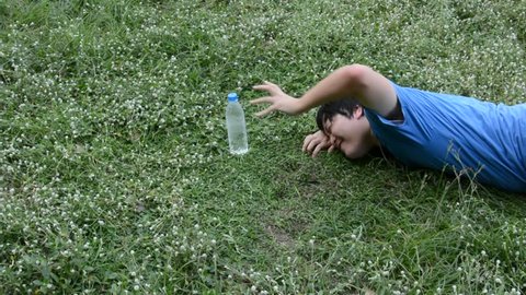 Asian Thai male guy is struggling crawling for a plastic drinking water bottle to survive his thirst and heatstroke on the empty green grass field but couldn't make it. It's a death concept.