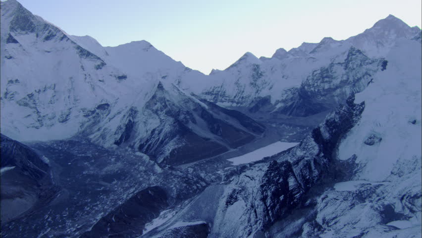 Snow Rocky Mountains Peaks Canyon. The shot shows a snow covered mountain range with sheer rock cliffs. Below a twisting bouldered canyon bed. The tall mountain ridges cast shadows over the canyon.