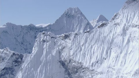White Snow Rocky Mountains. Steep mountain ridges covered in white snow extend to a major mountain peak. Shallow jagged rocks line and create a narrow side to the face of the mountain.