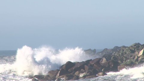 Large storm waves crashing on rocky inlet pier at Pacific Ocean with seagulls flying.