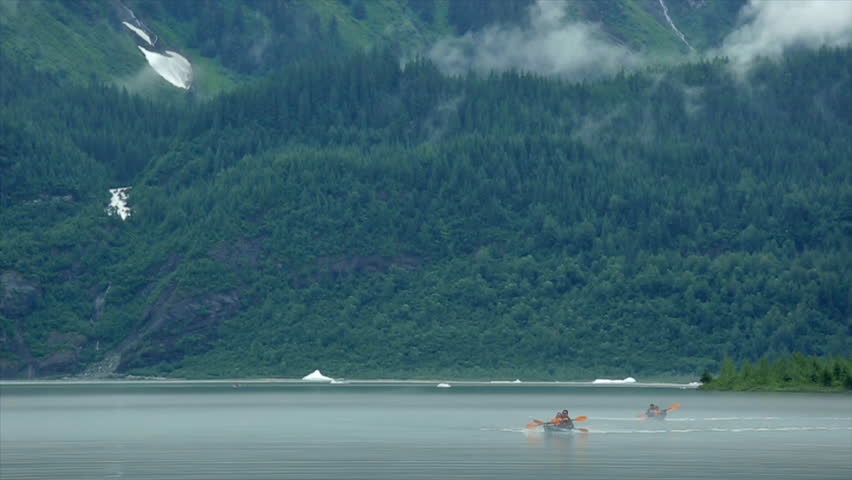 Two tandem kayaks paddle toward camera and away from foggy green mountains in Juneau, AK