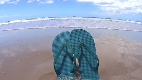 Wave washes in and knocks over a pair of flip flops stuck in the sand on the beach.