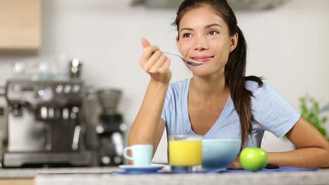 Woman eating breakfast cereals drinking orange juice smiling happy in the morning. Beautiful young multiracial woman sitting in her kitchen at home. Mixed race Asian Caucasian female model.