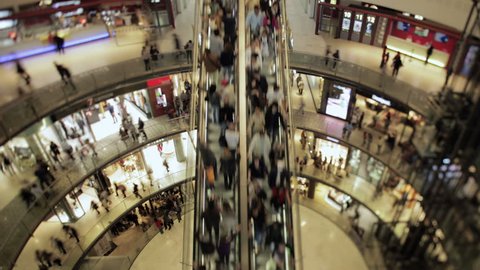 Shift and tilt wide angle time lapse of an escalator and people passing in a big round square at a mall. 4K