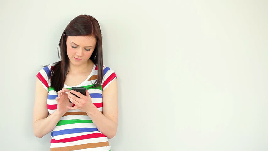 Smiling young woman texting on her phone against a white wall