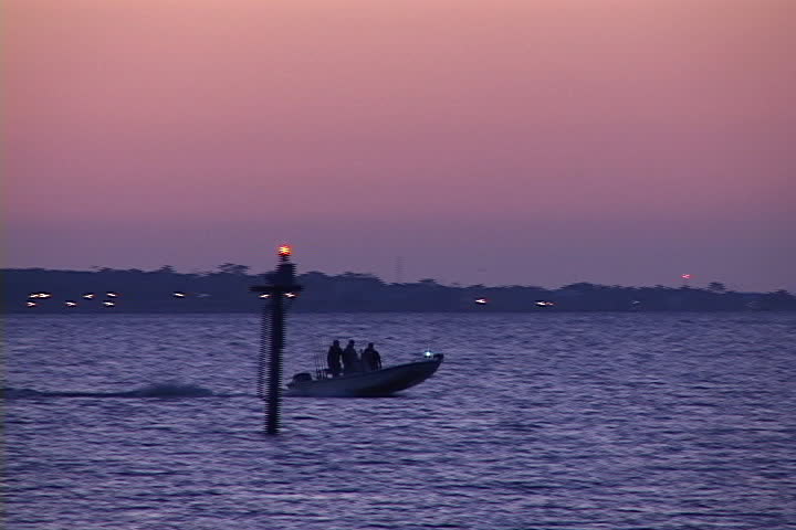 Fishermen speed a small fishing boat across the waters of lower Mobile Bay in Alabama at sunset with oil platforms and the Dauphin Island bridge in the background.