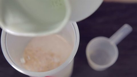 Man's hand tossing a scoop of chocolate whey isolate protein into plastic white shaker and pouring milk over the protein, with focus on the rim of the shaker 