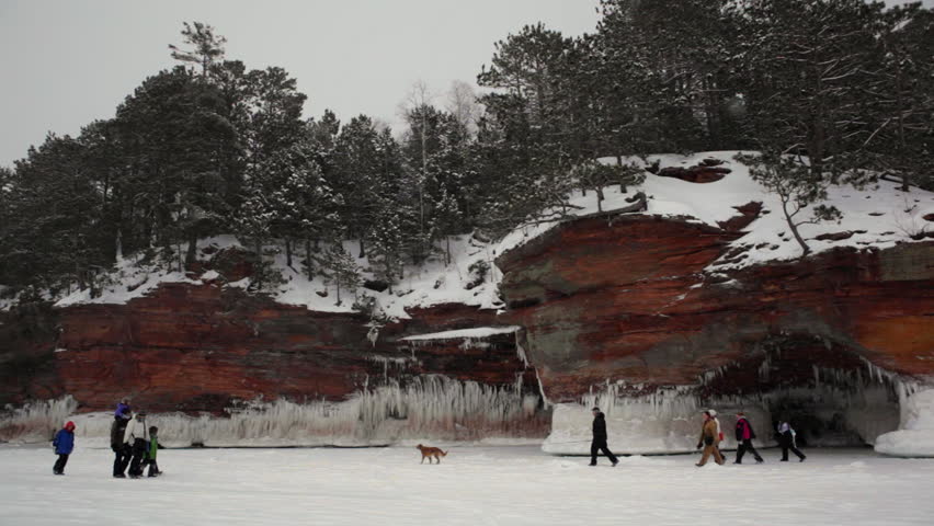 The Apostle Island mainland sea caves as ice caves on frozen Lake Superior. Hikers with children and dog passing in front of icy caves in huge snow-covered cliffs with forest above and falling snow.