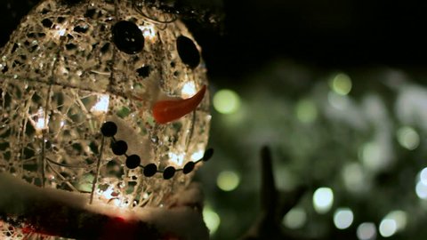 Close up dolly of an electric snowman, lit up at nighttime as it snows.