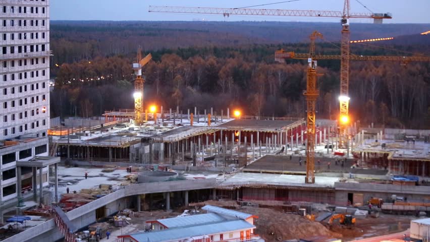 Construction site with buildings, moving cranes and workers in dusk.