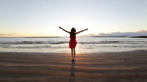 Beach woman running to water in dress at sunset raising arms enjoying freedom during summer holidays vacation travel. Beautiful happy free mixed race Asian Caucasian female girl outside.