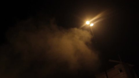 Smoke rises to the street light. Fog descended on the town. Smoke coming out from burning garbage container. Wind carries the smoke from a burning trash can. Horror, abstract background, night shot.