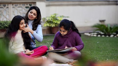 Shot of a woman combing her elder daughter's hair with younger one drawing in lawn Video Stok