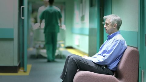 Time lapse. A worried man sits alone with his thoughts in a hospital waiting area as staff and other visitors rush around him.
