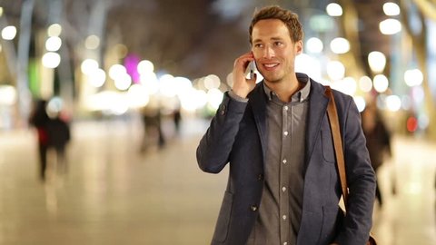 Casual young business talking on smart phone at night on La Rambla in Barcelona. Handsome young business man talking on smartphone and walking towards camera smiling happy wearing suit jacket outdoors