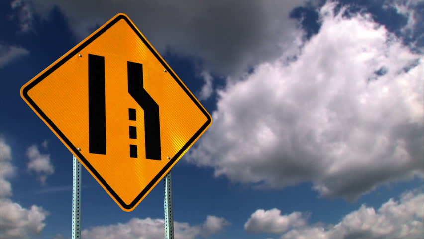 A merge ahead road sign with time lapse clouds.