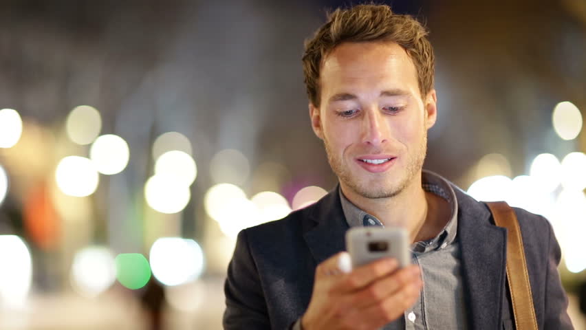 Man sms texting using app on smart phone at night in city. Handsome young business man using smartphone smiling happy wearing suit jacket outdoors. Urban male professional in his 20s.