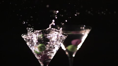 Colorful green and black olives on a cocktail stick are dropped in slow motion into a glass of martini, causing the drink to splash and spill. On a black background.