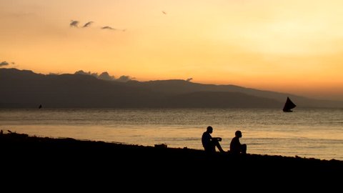 Port-au-Prince, Haiti - Jan 2009: Silhouetted people watch the Caribbean Sea at sunset on a beach, near Port-au-Prince.  Fishing boat sails behind them, in front of distant mountains.