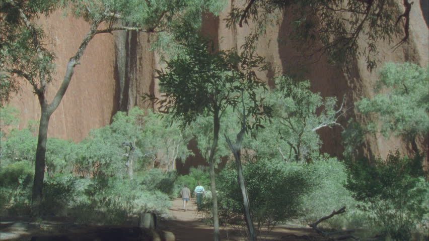tilt up from tourists on ground to perimeter of gorge with trees in foreground and rock of Kata Juta some blue sky behind