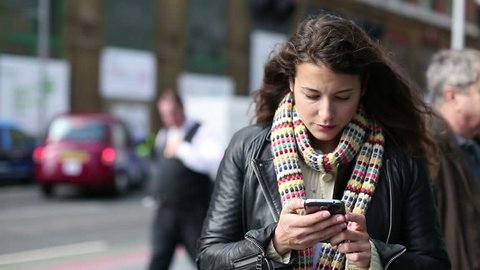 Attractive young woman using a smart phone and looking around whilst out on a public street