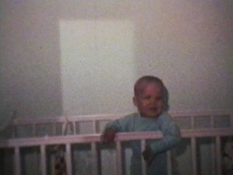 A cute little boy plays around in his crib and smiles for the camera.