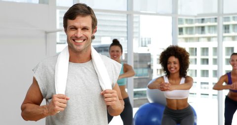 Fitness class squatting on bosu balls while instructor smiles at camera at the gym