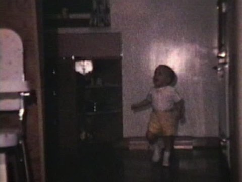 A cute little boy wearing yellow shorts walking around inside his house.