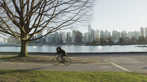 Dolly shot of cyclist riding along seawall at sunset with Vancouver skyline in background.

