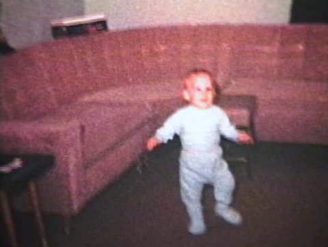 A little boy wearing a blue sleeper practices walking in the living room. (Vintage 8mm film)