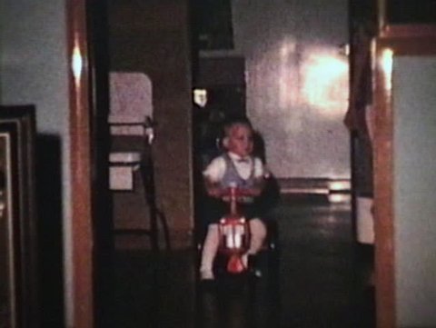 A cute little boy rides his toy tractor around the house and then crashes while being pushed backwards. (Vintage 8mm film)